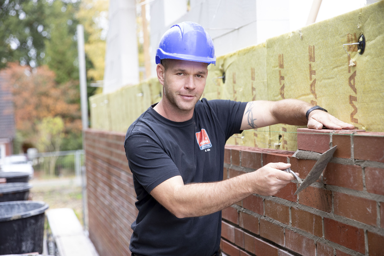 Maurer vor einer Backsteinmauer, mit Spachtel in der Hand und blauem Helm auf dem Kopf